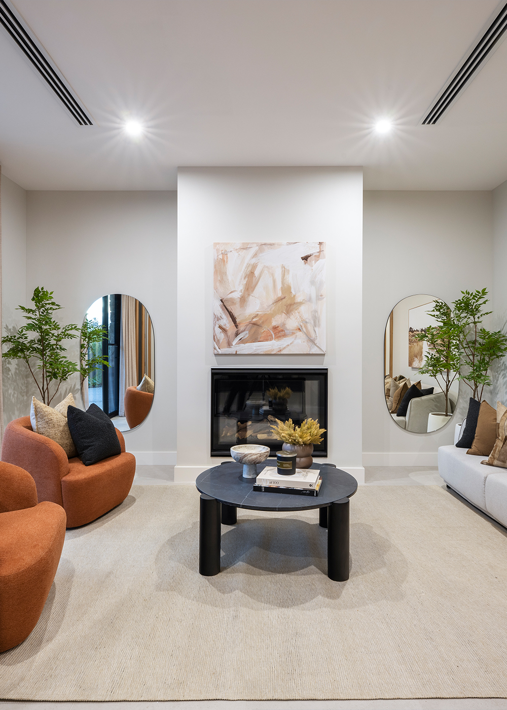 Front sitting room with gas fireplace, a pair of curved armchairs in a burnt orange fabric sit beside the windows, opposite a 3 seater grey lounge suite.