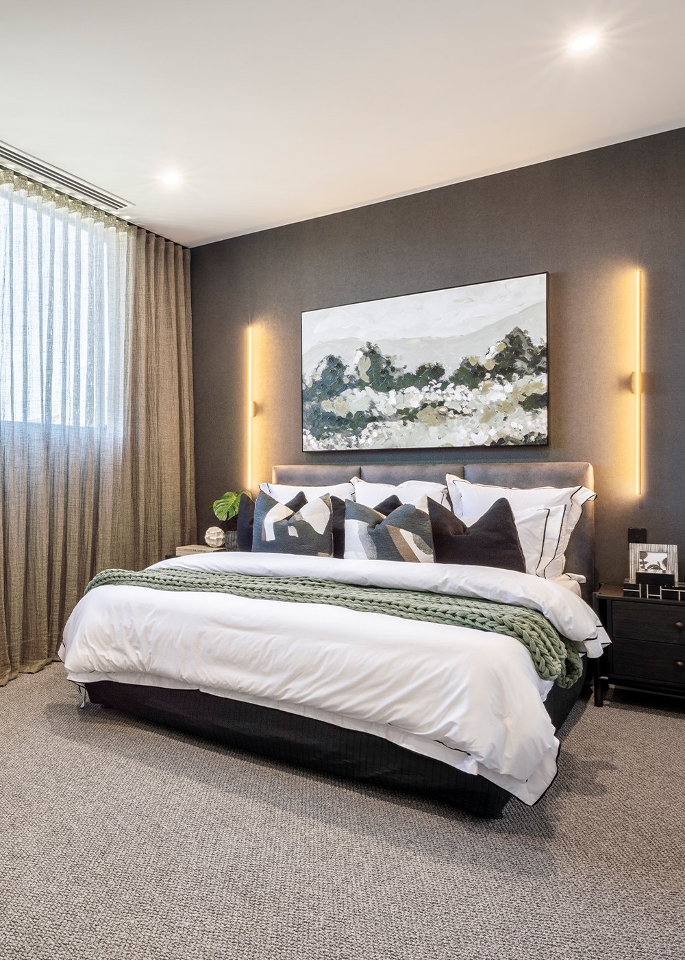 Main bedroom with king size bedding dressed with white and dark accent pillows and soft green throw. Back wall is decorated with wallpaper and up lighting