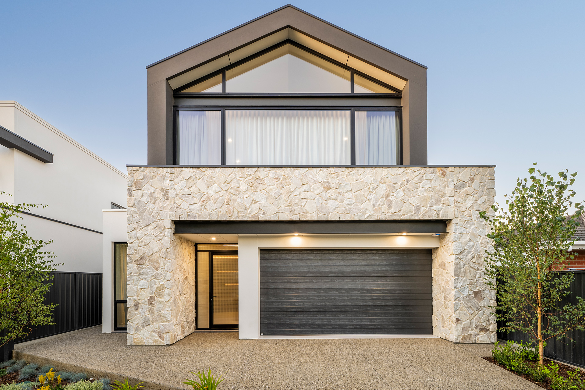 Grand prize home facade with stone wall surrounding the entry and garage, upper level features a modern peak roof line. Some garden appears to each side and clear blue skies above